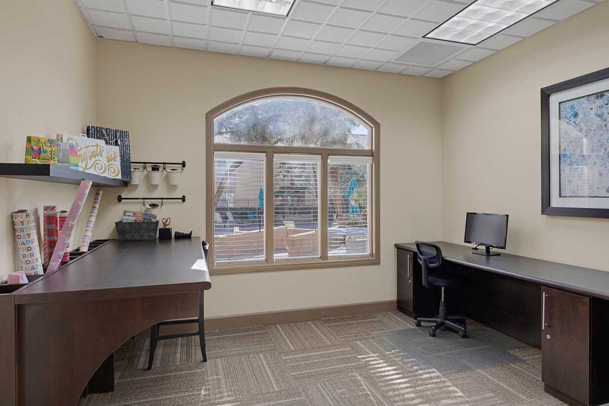 A well-lit office space featuring two desks, one with a computer, shelves displaying colorful items, and a large window with a view of an outdoor area. The walls are painted a neutral color, and the flooring is a patterned tile, creating a clean and organized atmosphere.