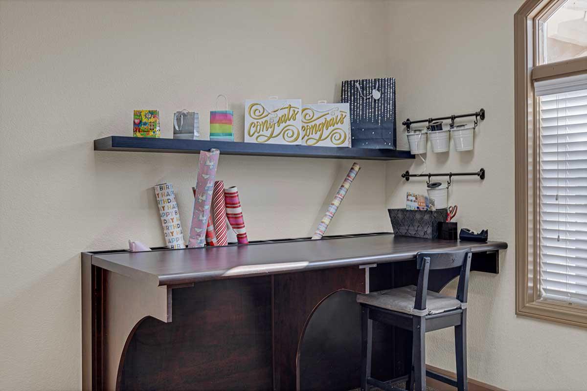 A neatly organized workspace featuring a dark wooden desk against a light wall. There are colorful gift-wrapping rolls, decorative items on the shelf, and a "Congrats" sign. A chair is positioned at the desk, with clips and containers for supplies hanging above. Natural light streams in from the window.