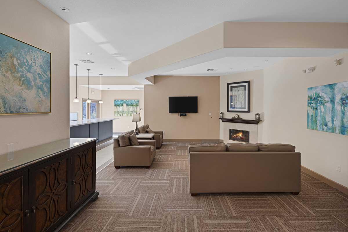 A modern living room with two beige sofas, a flat-screen TV mounted on the wall, and a decorative fireplace. The room features two abstract artworks on the walls and a wooden cabinet. Large windows allow natural light to illuminate the space, which has neutral-colored walls and a patterned carpet.
