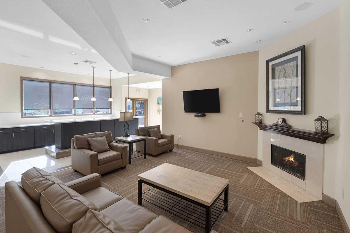 A cozy living room featuring two brown leather armchairs and a matching sofa, arranged around a coffee table. A modern flat-screen TV is mounted on the wall, and a gas fireplace adds warmth. The space is well-lit with large windows and a sleek kitchen visible in the background.