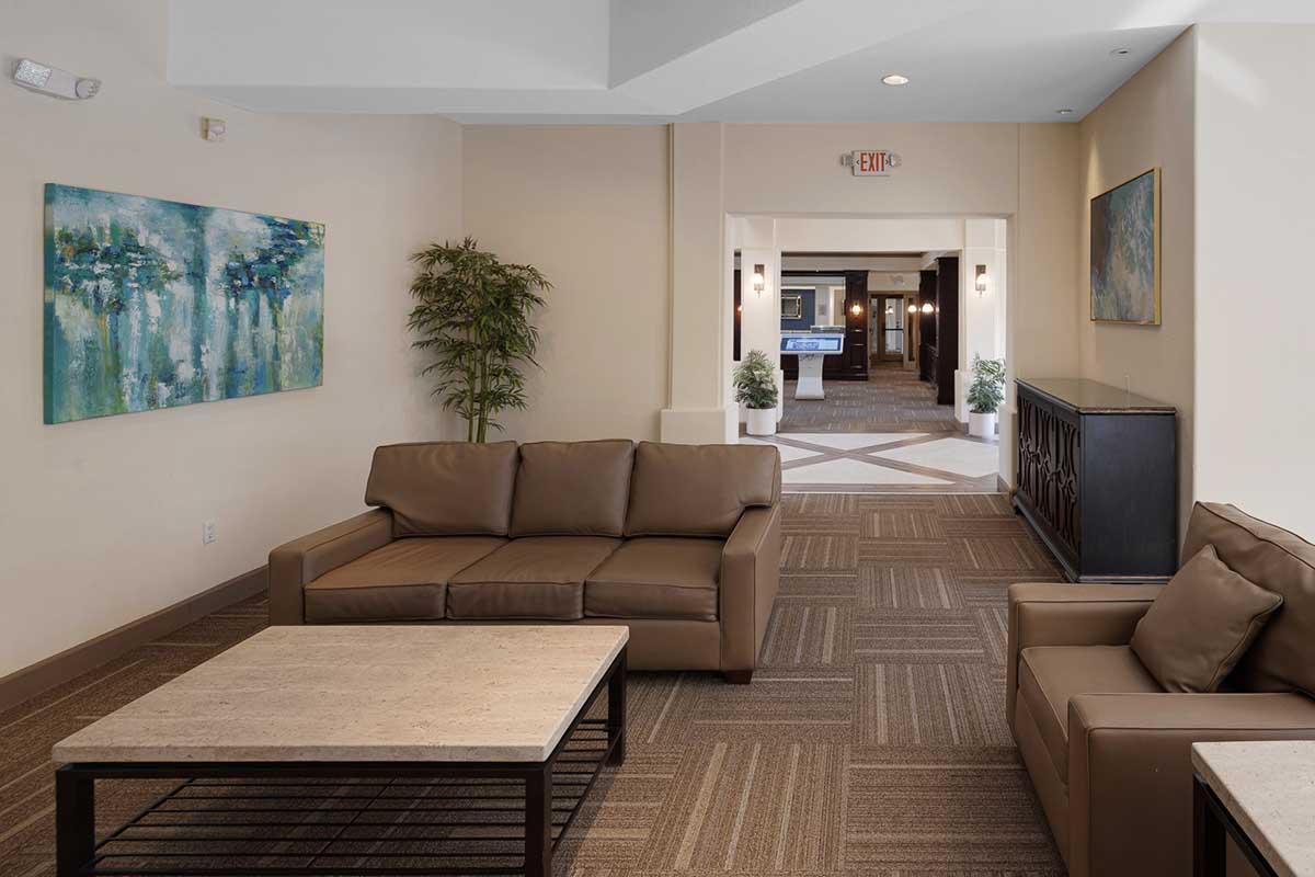 A modern lobby area featuring a brown leather sofa and an armchair with a marble coffee table. The walls are painted in neutral tones, adorned with a blue abstract painting. A potted plant adds a touch of greenery, and an exit door is visible in the background. The flooring consists of textured carpet in earthy tones.