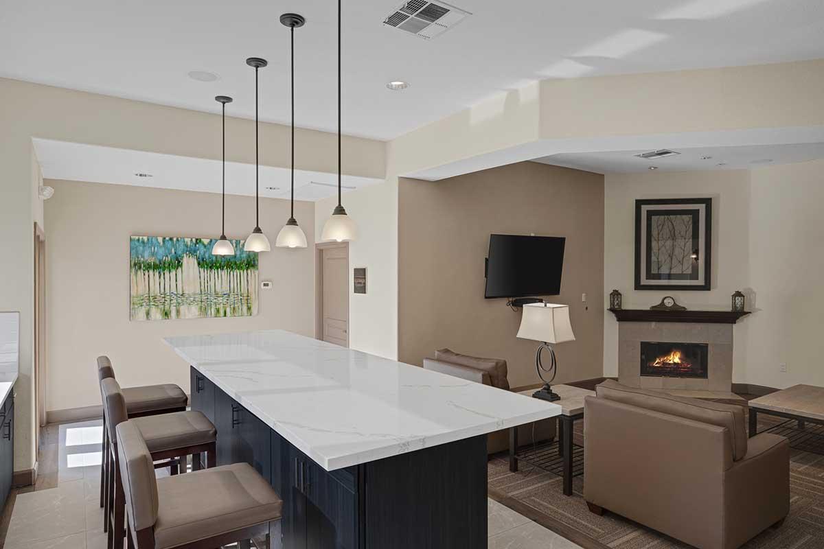 A modern living room with a spacious bar area featuring a white marble countertop. There are four tall bar stools, two comfortable brown leather chairs, and a television mounted on the wall. A cozy fireplace is present, and a piece of artwork depicting green trees hangs above the seating area.