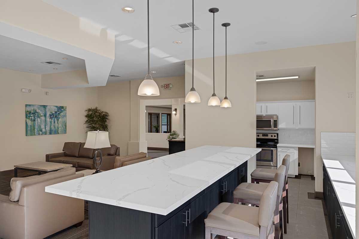 A modern kitchen and dining area featuring a long white marble countertop with bar seating, pendant lights hanging above, a sofa and armchairs in a cozy living space, and a well-equipped kitchen with white cabinets and stainless steel appliances. Soft, neutral colors create a welcoming atmosphere.