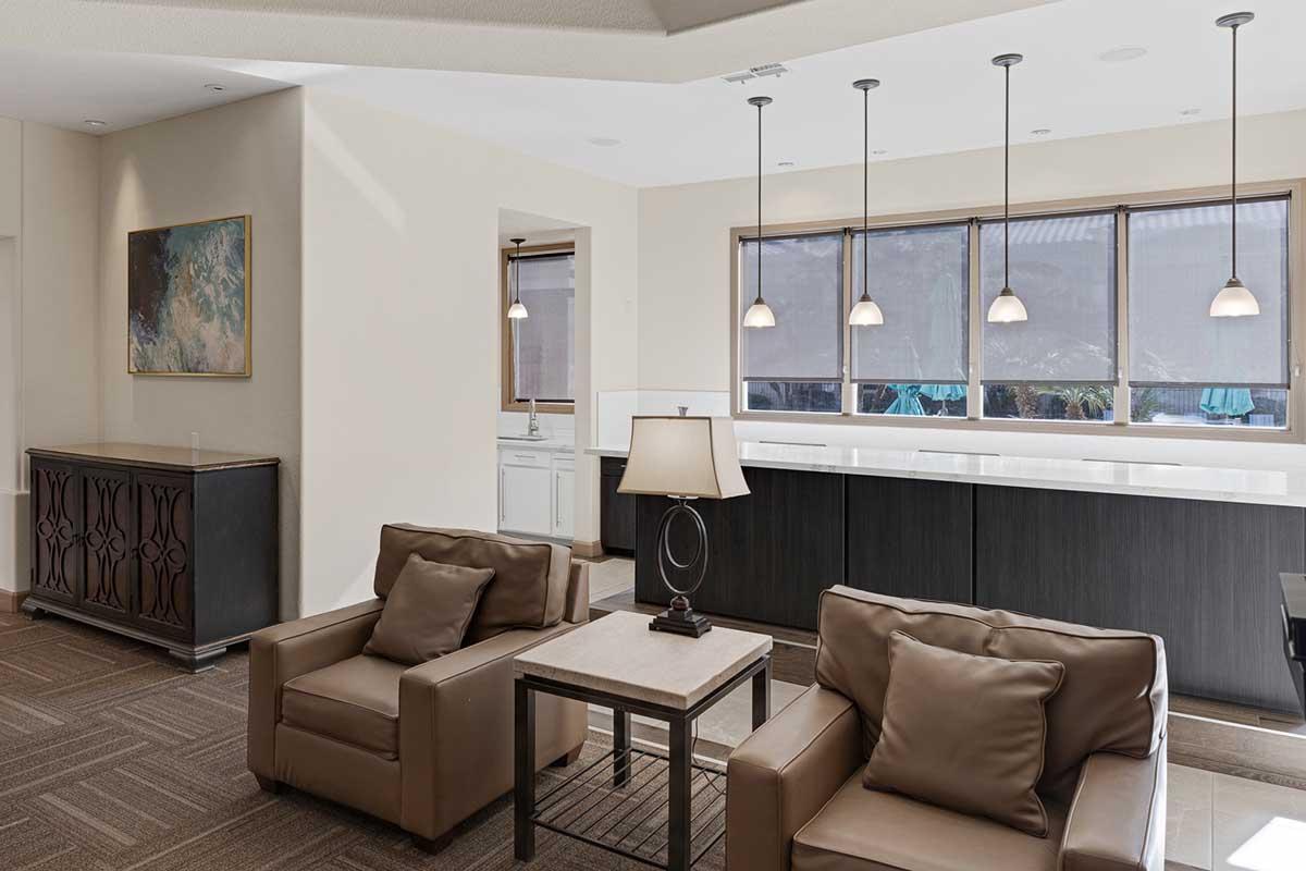 A modern, brightly lit lounge area featuring two brown leather chairs with cushions, a small coffee table, and a lamp. In the background, there is a sleek bar with pendant lighting and large windows showing an outdoor view. The decor is minimalist and contemporary.