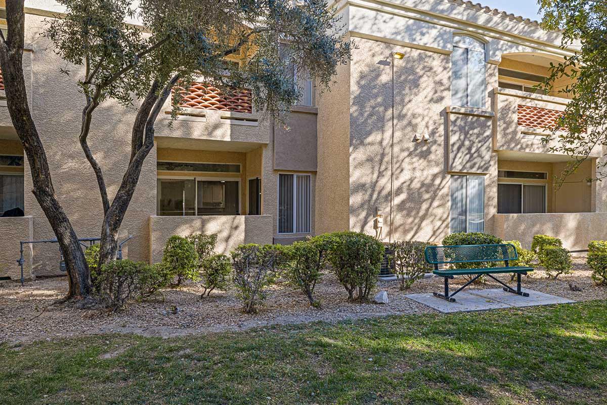 A view of an outdoor area featuring a well-maintained grassy space with a bench, surrounded by small shrubs and trees. In the background, there are two tan buildings with balconies and large windows. The scene is bright and inviting, suggesting a peaceful residential setting.