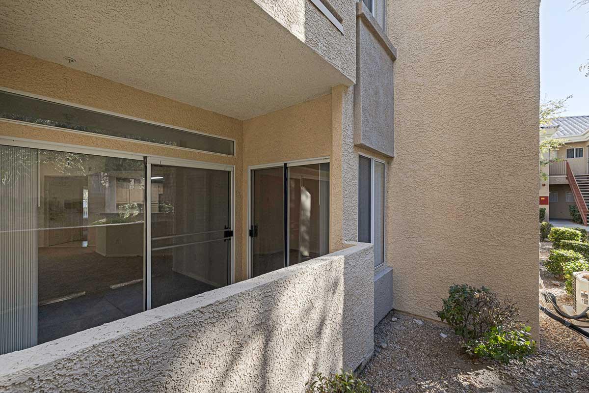 A corner view of a beige apartment building, featuring a covered patio area with sliding glass doors. The surrounding landscape includes small shrubs and a clear path. Natural light filters through the windows, indicating a well-lit space.