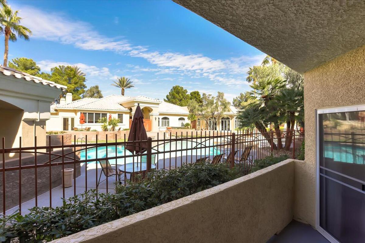 View from a balcony overlooking a serene pool area surrounded by palm trees and landscaped bushes. Sunlight brightens the scene, showcasing a well-maintained community with lounge chairs and umbrellas near the pool, creating a relaxing atmosphere.