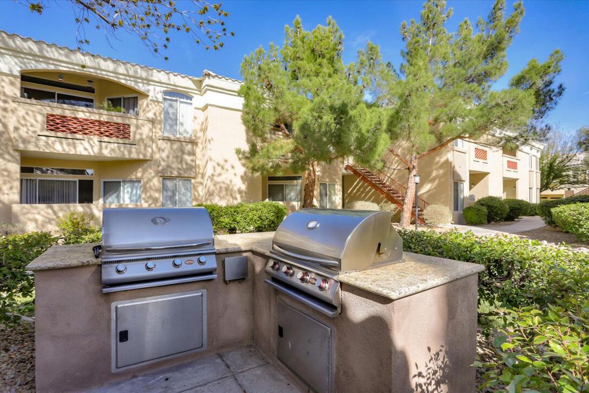 Outdoor barbecue area featuring two stainless steel grills set against a backdrop of landscaped greenery and residential buildings. The space is surrounded by low shrubs and trees, with clear blue skies overhead, creating a pleasant atmosphere for outdoor cooking and gatherings.