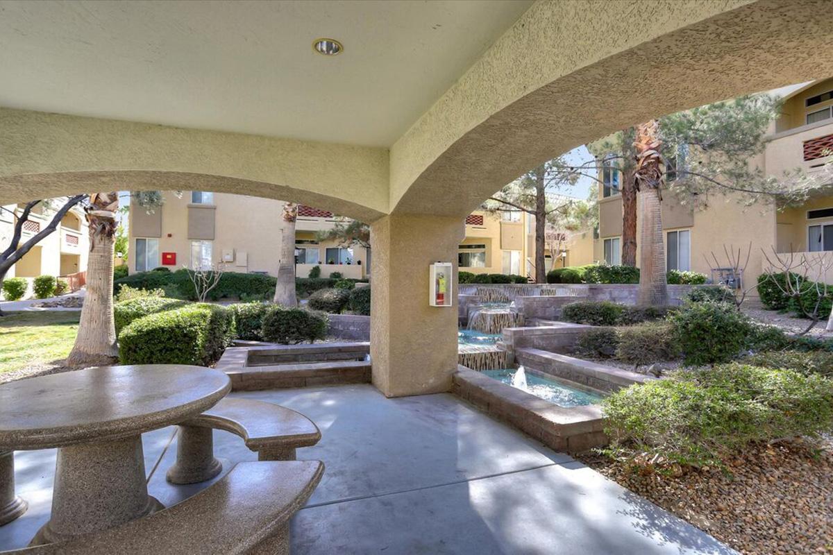 A serene outdoor area featuring a stone picnic table with benches under an archway, surrounded by lush greenery and small trees. In the background, a decorative water feature adds to the peaceful atmosphere next to residential buildings. Bright sunny day enhances the inviting scenery.