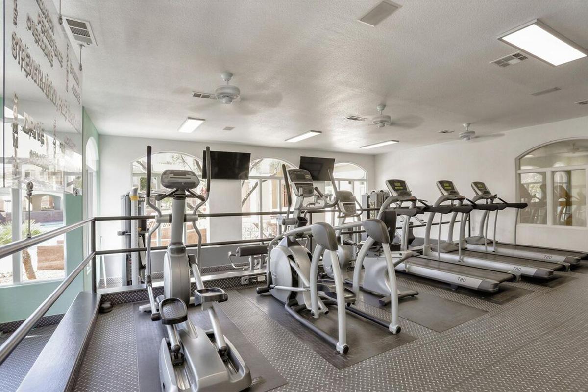 A well-lit fitness room featuring several elliptical machines and treadmills facing a large window. The walls are painted light colors, and there are wall-mounted televisions. The space appears clean and modern, with a rubber mat flooring, suitable for workouts.