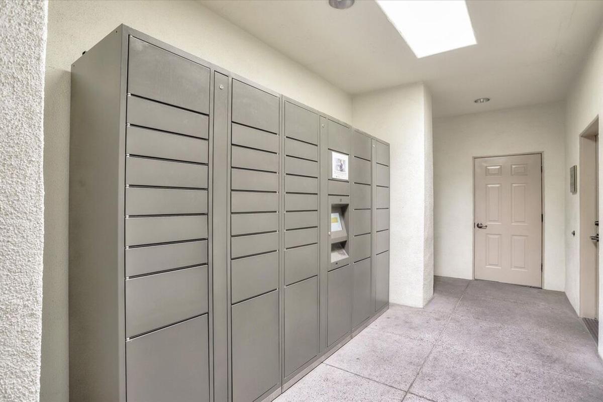 A row of metallic mail lockers installed in a hallway, with a nearby door. The lockers are gray and feature multiple compartments for package deliveries. The space is well-lit, with a skylight above, and the floor is a light-colored concrete.