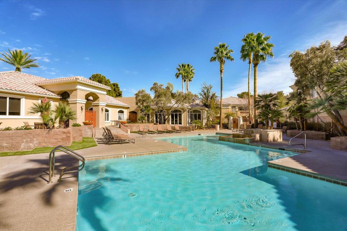A serene outdoor swimming pool surrounded by palm trees and lounge chairs. The pool area features a spacious deck with light blue water under a clear blue sky, and a stylish building with a Mediterranean design in the background. Ideal for relaxation and leisure.