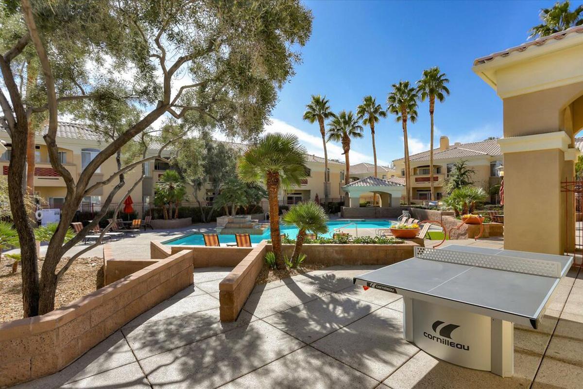 A bright outdoor scene featuring a swimming pool surrounded by palm trees and lounge chairs. A ping pong table is positioned on a tiled patio, with a few buildings visible in the background. The sky is clear and blue, suggesting a warm, sunny day.