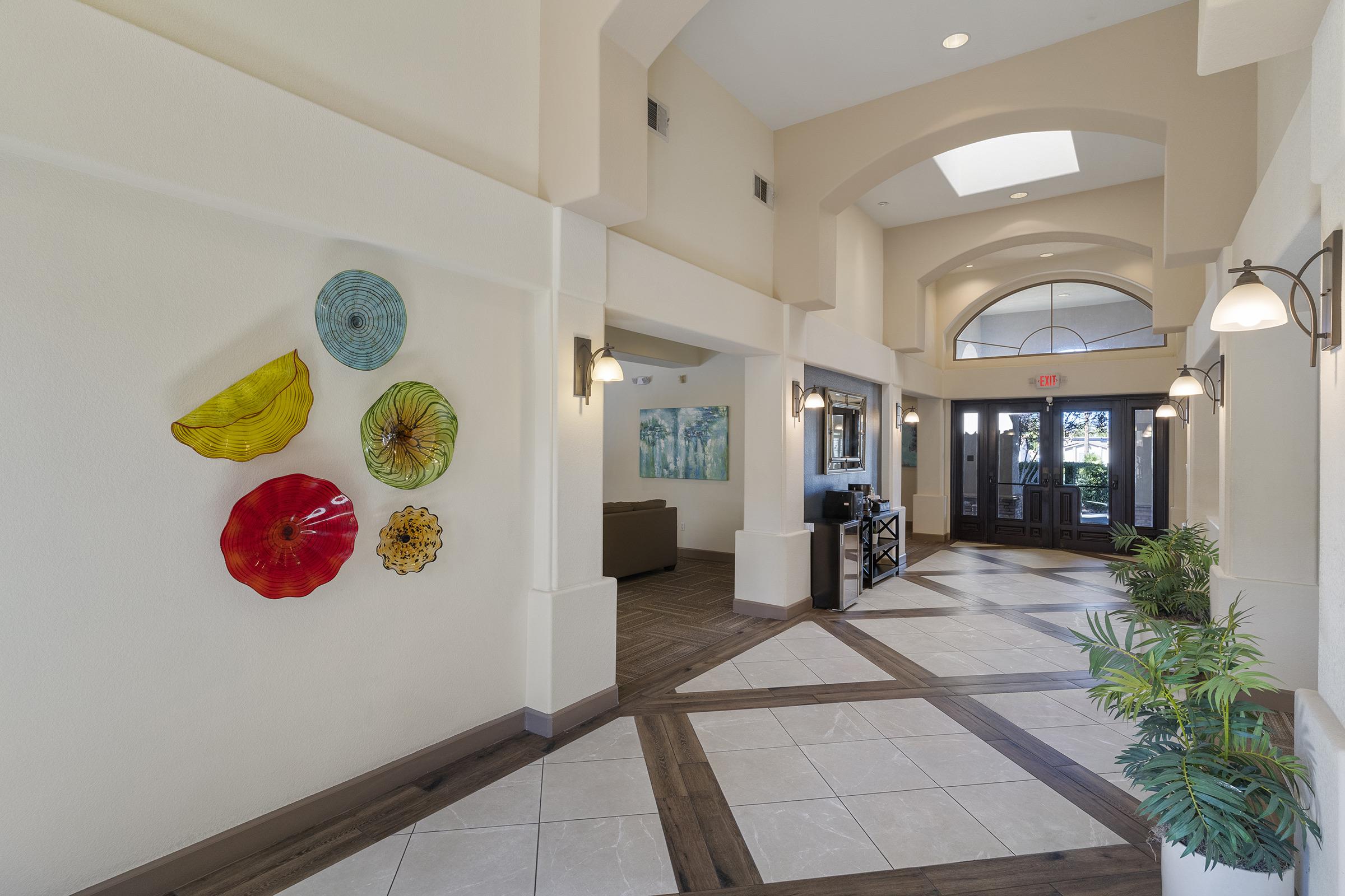 An interior view of a spacious lobby featuring a decorative wall with colorful glass art pieces in various shapes. The floor is made of tiled patterns with wooden accents. There are lounge seating areas visible, along with large windows allowing natural light to illuminate the space. Plants are placed throughout for added decor.
