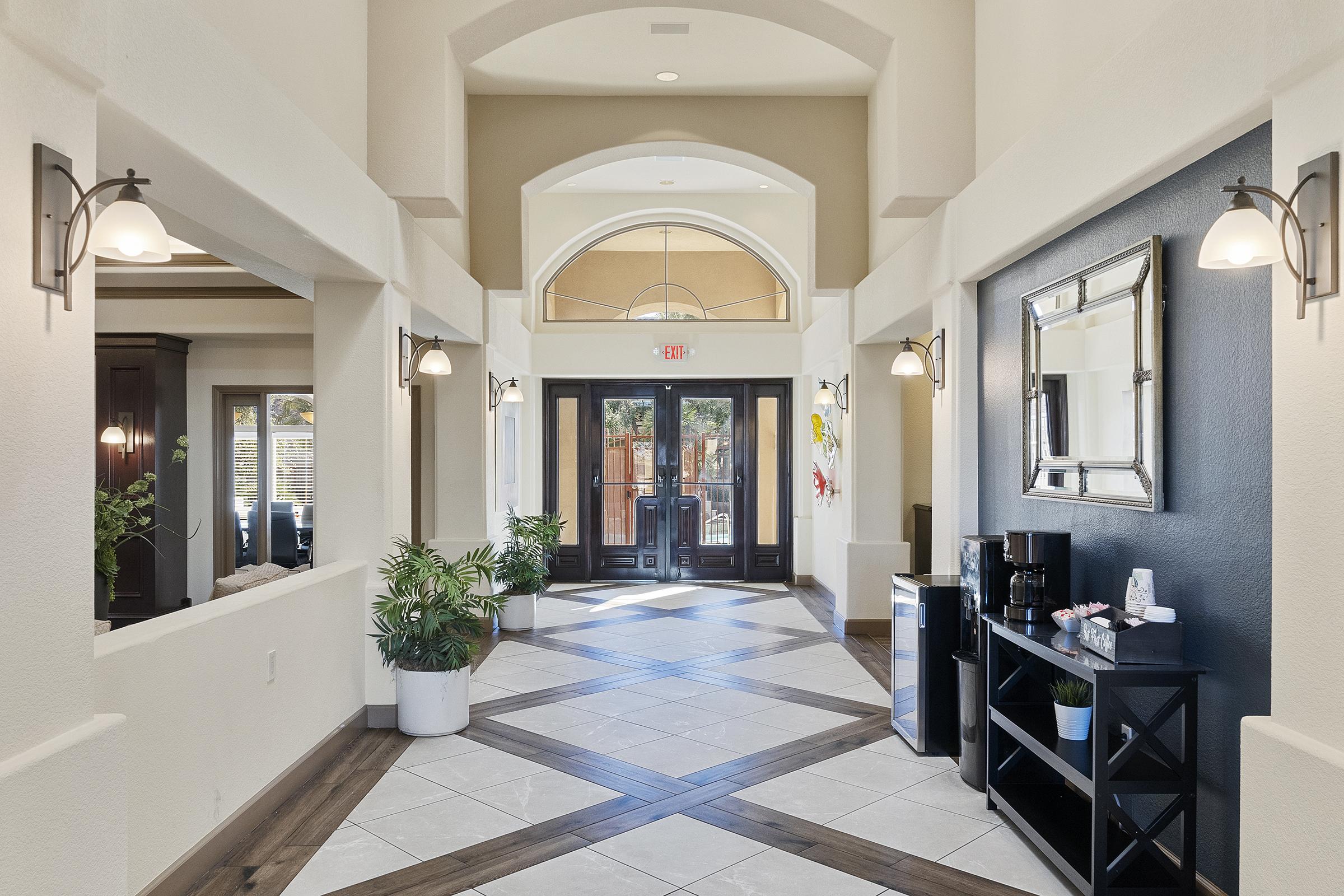 A well-lit hallway featuring glossy floor tiles and textured wall panels. Decorative lighting fixtures are mounted on the walls, and there are potted plants on display. At the far end, double doors open to an outdoor area, and a dark wooden console with a coffee station is positioned on one side.
