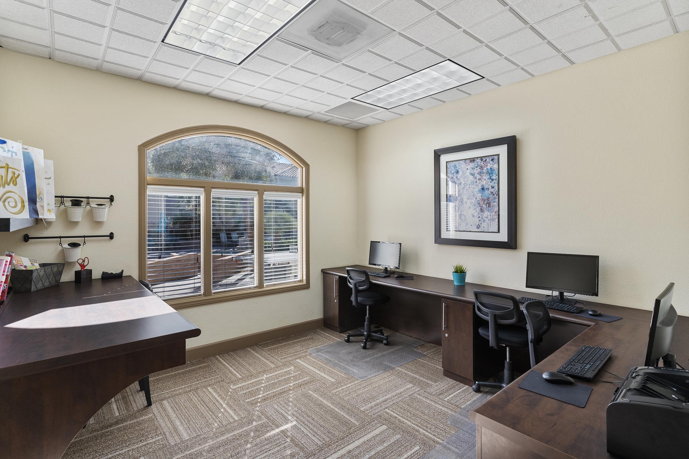 A bright, modern office space featuring two desks with computers, a large window with a view outside, and neutral-colored walls. The room has a stylish framed artwork, a small potted plant, and organized desk items, creating a comfortable and productive environment.