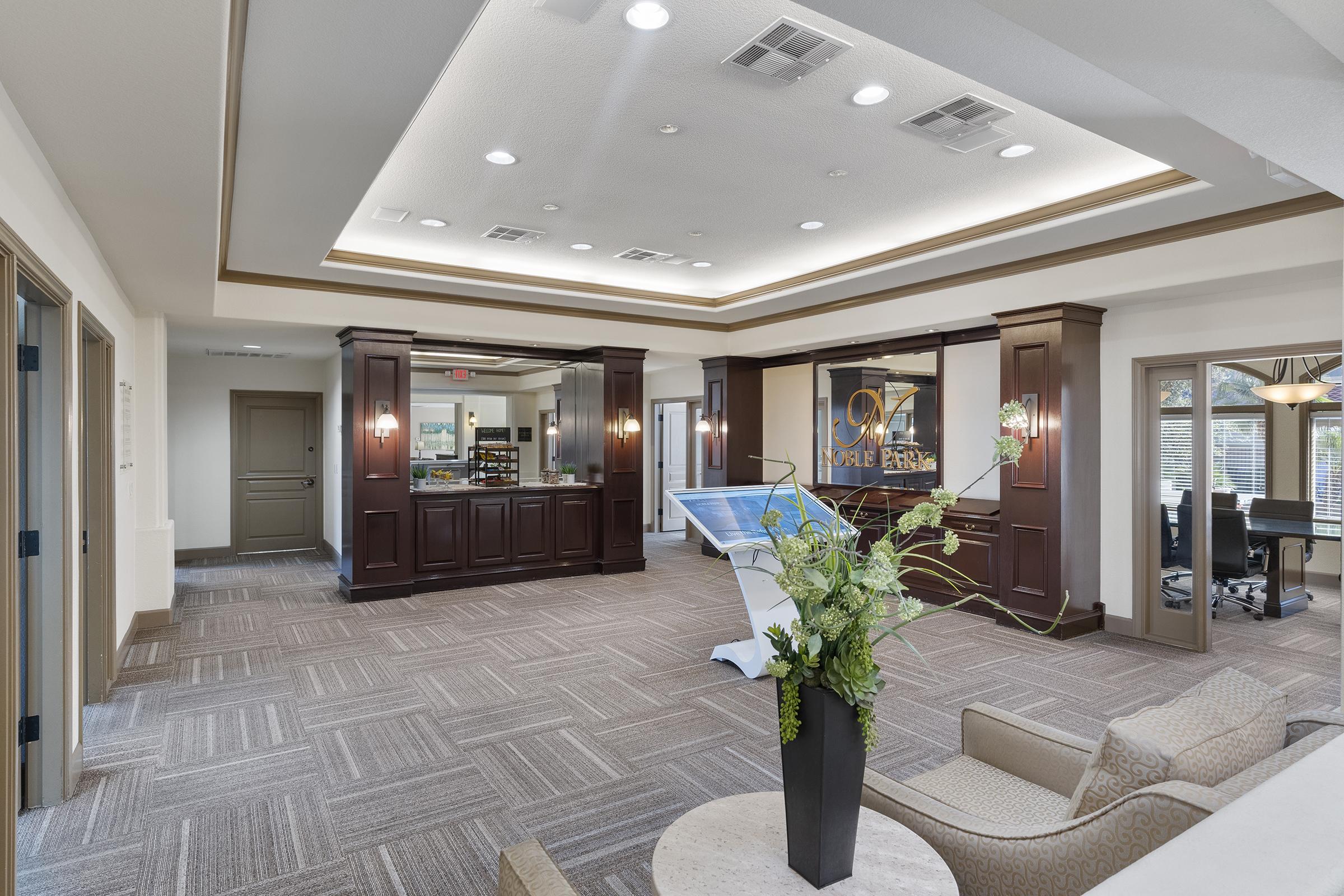A modern reception area with a spacious layout featuring a decorative seating area, a front desk made of dark wood, and a large informational display. The room is well-lit with overhead lights and has stylish decor elements, including a potted plant and elegant finishes.