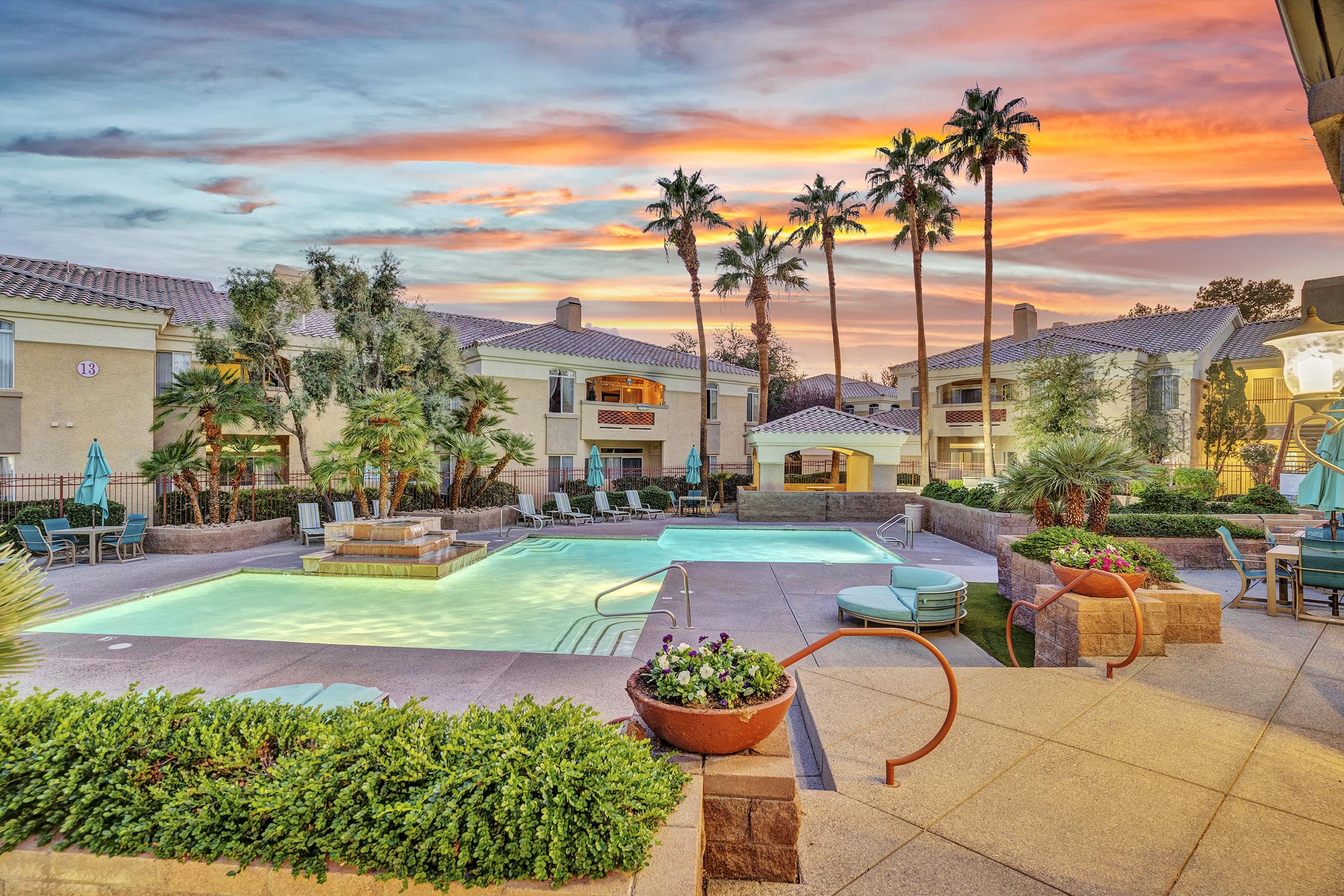 A vibrant sunset over a resort-style pool area surrounded by palm trees and lush landscaping. The pool, illuminated with a gentle glow, features a small fountain and is bordered by comfortable lounge chairs and tables. The buildings in the background add to the relaxing atmosphere.