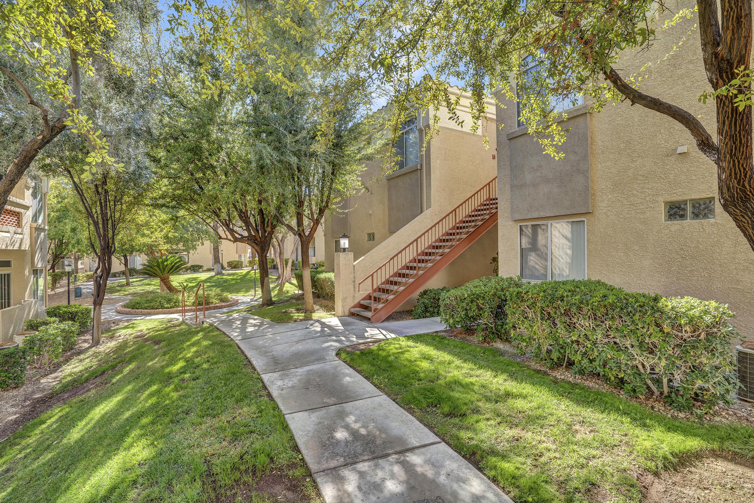 A well-maintained walkway surrounded by lush green landscaping, leading to an apartment complex. There are trees providing shade, and a set of stairs leading to upper-level units. The scene is bright and inviting, showcasing a residential atmosphere.