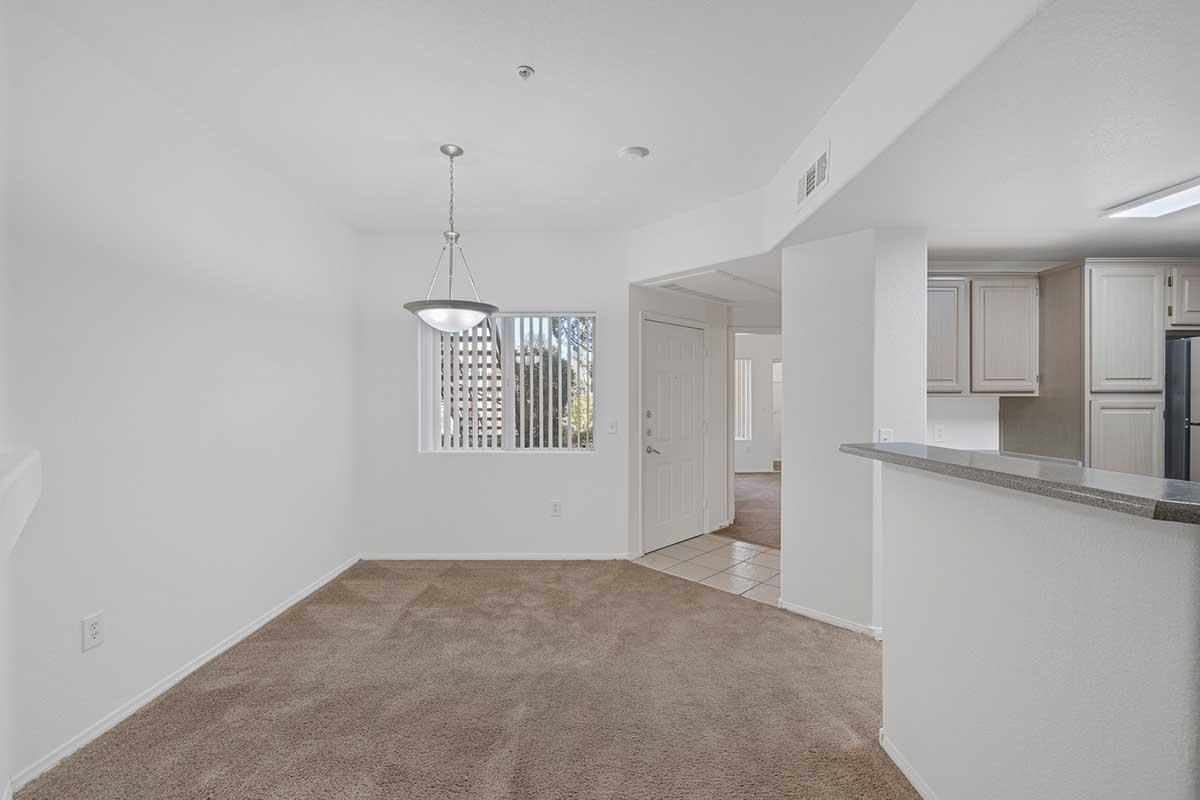 A spacious, well-lit interior of an apartment featuring beige carpet flooring, a window with vertical blinds, and a modern light fixture hanging from the ceiling. The open layout includes a kitchen area visible in the background with light-colored cabinetry and tile flooring.
