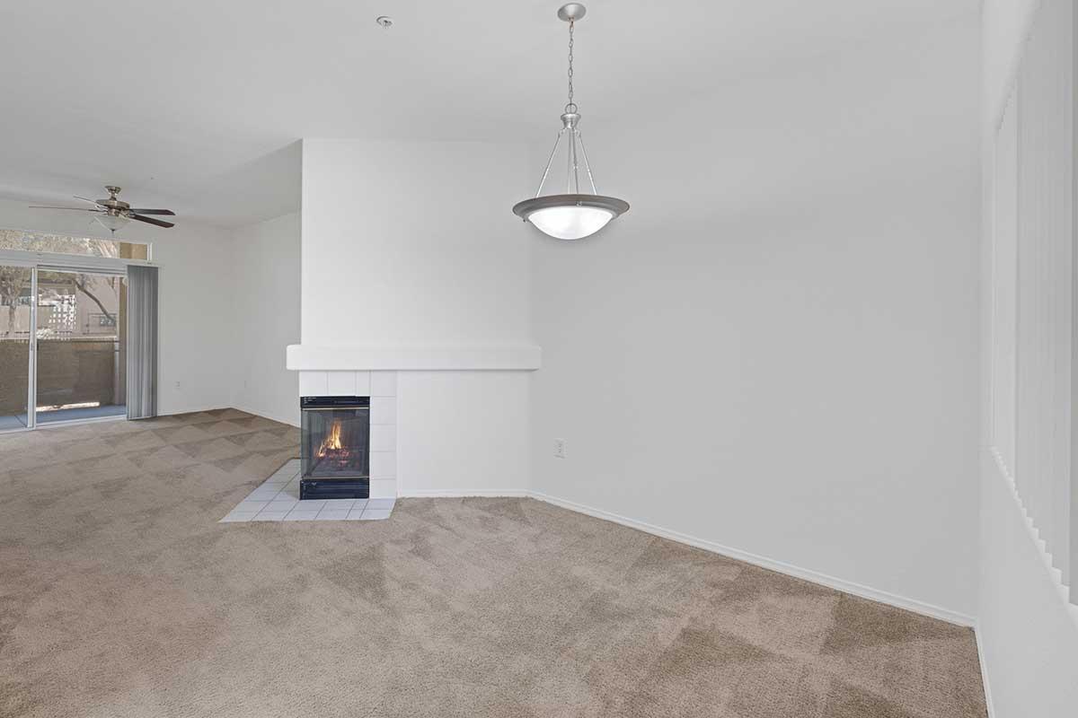 Spacious living room with beige carpet and a white wall, featuring a cozy fireplace with a blue tile surround. A ceiling fan is installed, and a pendant light hangs from the ceiling. Large windows allow natural light in, with glass doors leading to a balcony area. The overall atmosphere is inviting and warm.