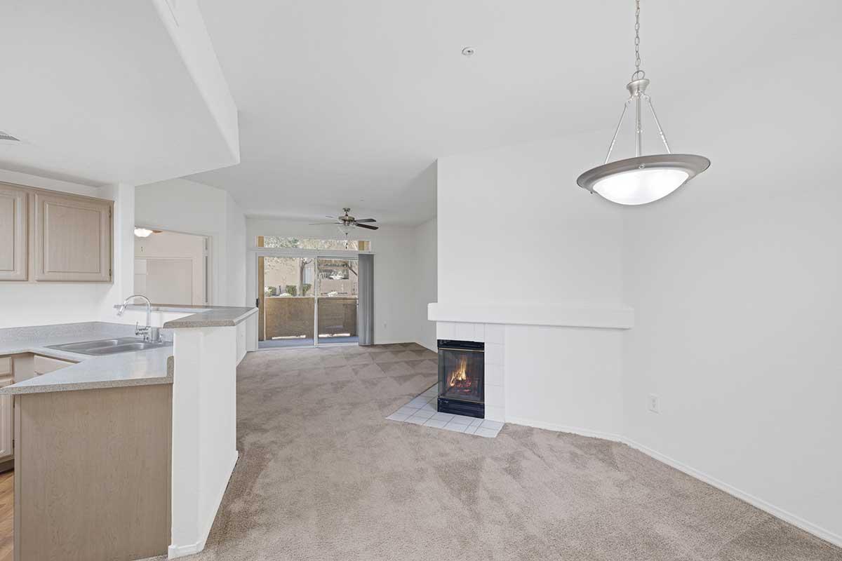 Bright, open living space featuring light-colored walls and carpet, a cozy fireplace with a tile surround, and a chandelier hanging from the ceiling. The kitchen is visible to the left with wooden cabinets, and sliding glass doors lead to a balcony or patio area.