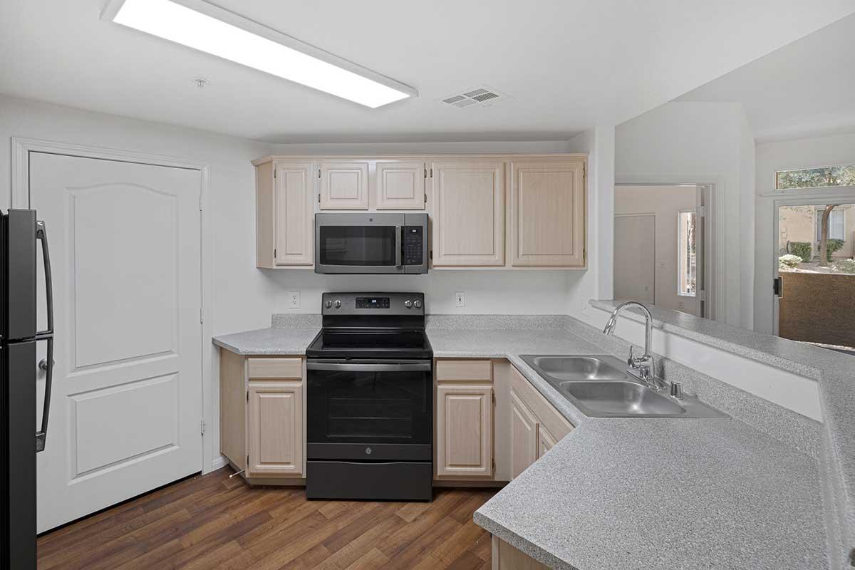Modern kitchen featuring light wood cabinets, a gray countertop, a black stove and oven, a microwave, and dual sinks. The space is well-lit with a ceiling light and includes a refrigerator on the side, with an open layout leading to a doorway and window.