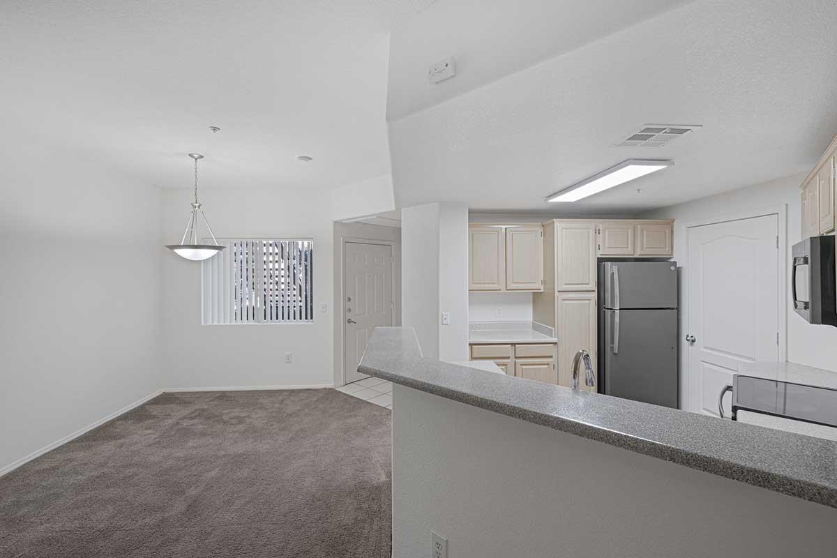 A spacious, well-lit living area with beige carpeting and white walls. There is a dining area with a light fixture, a kitchen bar with a gray countertop, and a modern kitchen featuring stainless steel appliances. A window allows natural light to enter, enhancing the bright and inviting atmosphere.