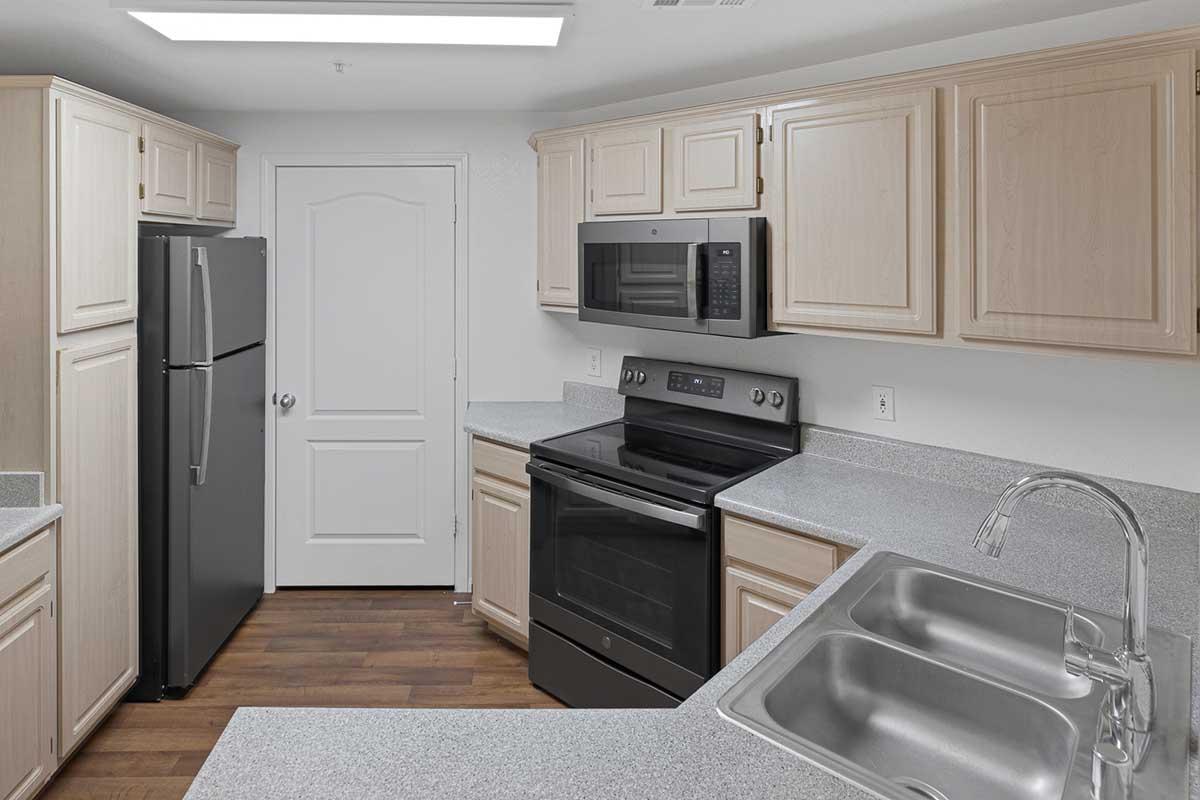 Modern kitchen featuring light wood cabinetry, stainless steel appliances including a refrigerator and microwave, a black oven, and a double sink. Countertops are gray and the flooring is a warm wood tone. A door leads to another room, and there is ample space for cooking and preparation.