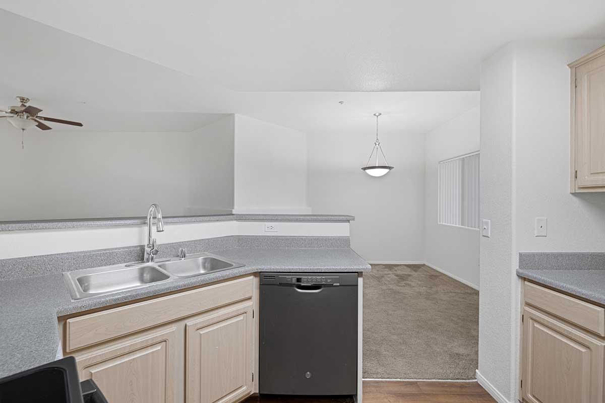 A modern kitchen featuring light wooden cabinets, a stainless steel dishwasher, and a double sink. The countertop is grey, and there is an open layout leading into a spacious living area with carpeted flooring and a ceiling light fixture. A large window allows natural light into the room.
