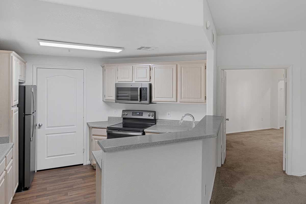 A modern kitchen with light wood cabinetry and gray countertops. It features a black refrigerator, a stainless steel microwave above a stove, and an open layout leading to a carpeted area in an adjoining room. The walls are painted a light color, enhancing the bright and spacious feel.