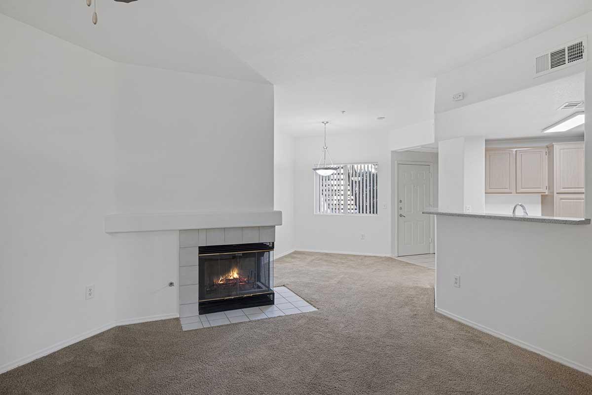 A spacious living area with beige carpeting, featuring a white fireplace with a tiled hearth. There is a large window allowing natural light, and an open kitchen area with light-colored cabinets visible in the background. The walls are painted white, creating a bright and airy atmosphere.
