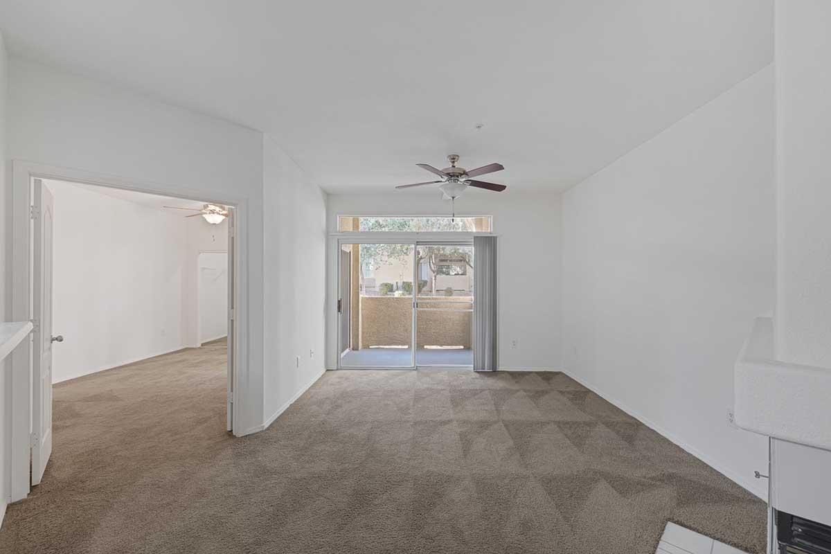 A spacious living room with beige carpet, light-colored walls, and a ceiling fan. The room features large sliding glass doors that lead to a balcony. To the left, there is an entrance to another room, and the overall ambiance is bright and airy.