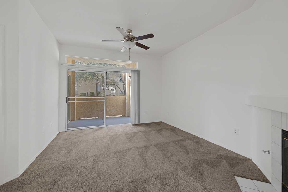 Empty living room with beige carpet and white walls. A ceiling fan hangs from the ceiling, and a sliding glass door leads to a balcony. Natural light enters through the door, illuminating the room. There is a fireplace in the corner, enhancing the cozy atmosphere.