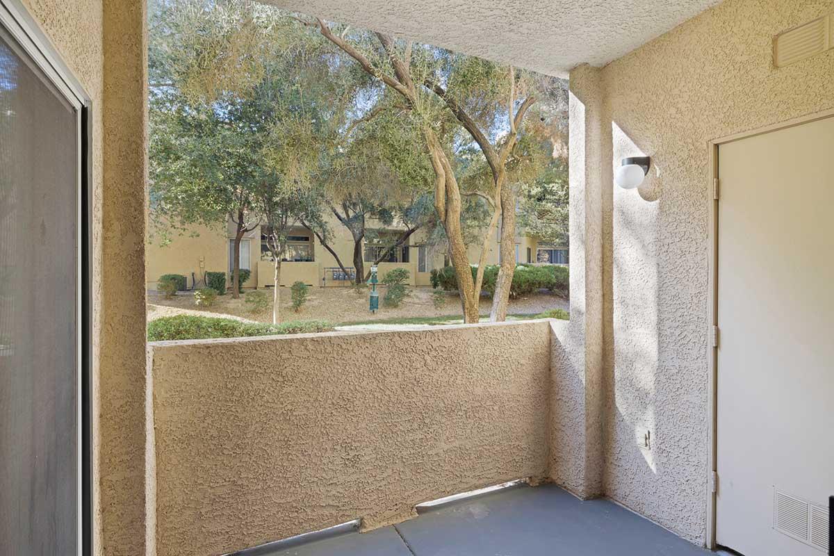 A view from a balcony overlooking a landscaped area with trees and shrubs. In the background, there are buildings and a walkway visible through the greenery. The balcony features a textured wall and a door, providing a cozy outdoor space.