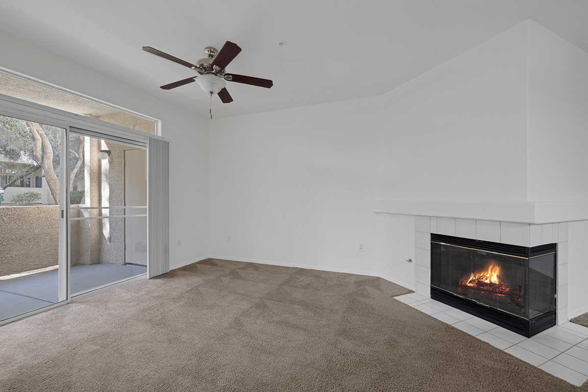 A spacious, empty living room with light beige carpet, featuring large sliding glass doors leading to a balcony. The room has a white ceiling fan and a modern fireplace with a white tile surround, adding a cozy touch to the neutral decor. Natural light fills the space.
