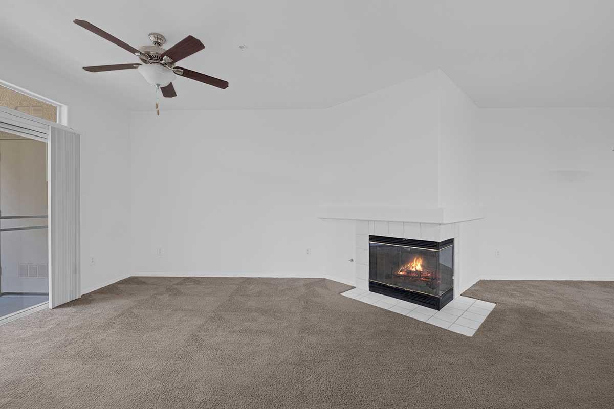 A spacious living room featuring a ceiling fan, light-colored walls, and a cozy fireplace with a tiled hearth. The floor is covered in brown carpet, and there is an open entry to a terrace or balcony on one side. The overall ambiance is bright and airy, suitable for relaxation and gathering.