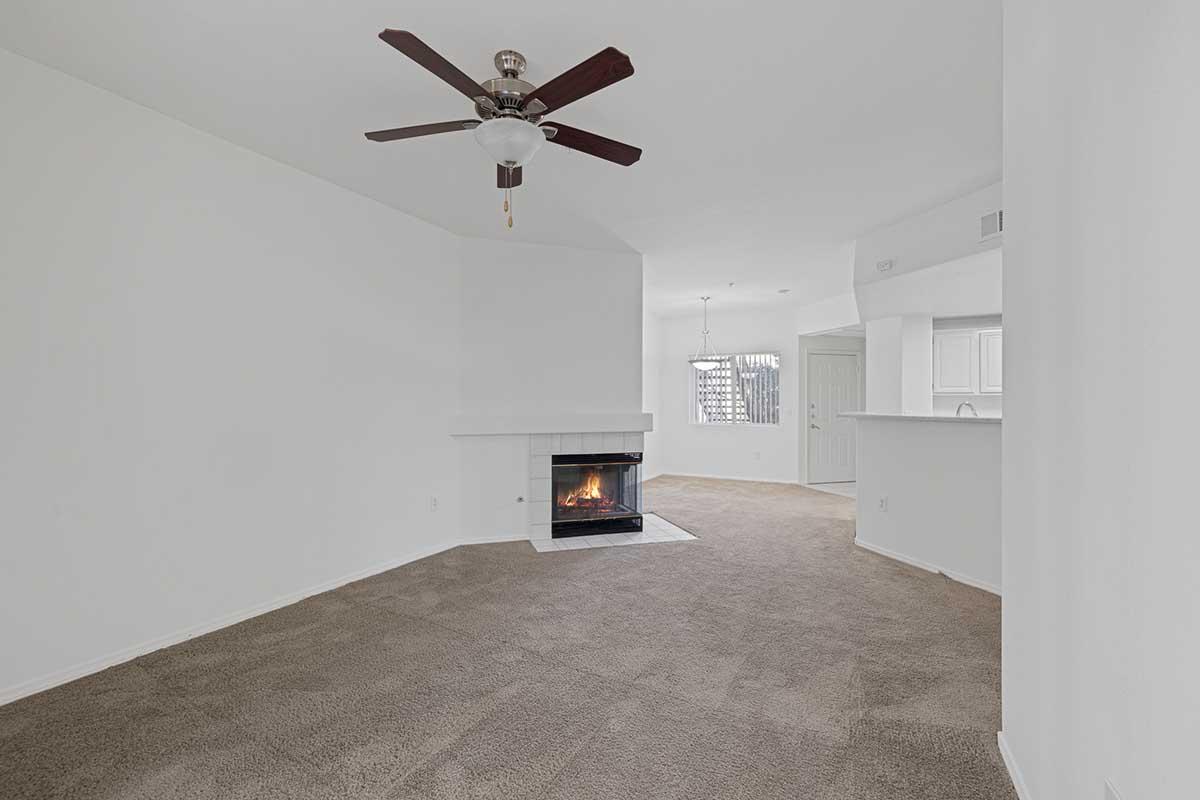 Interior view of a spacious living room featuring a ceiling fan, a fireplace with a black hearth, and light-colored walls. The room has a cozy carpeted floor and an open layout that leads to another area, possibly a kitchen or dining space, with natural light coming through a window.