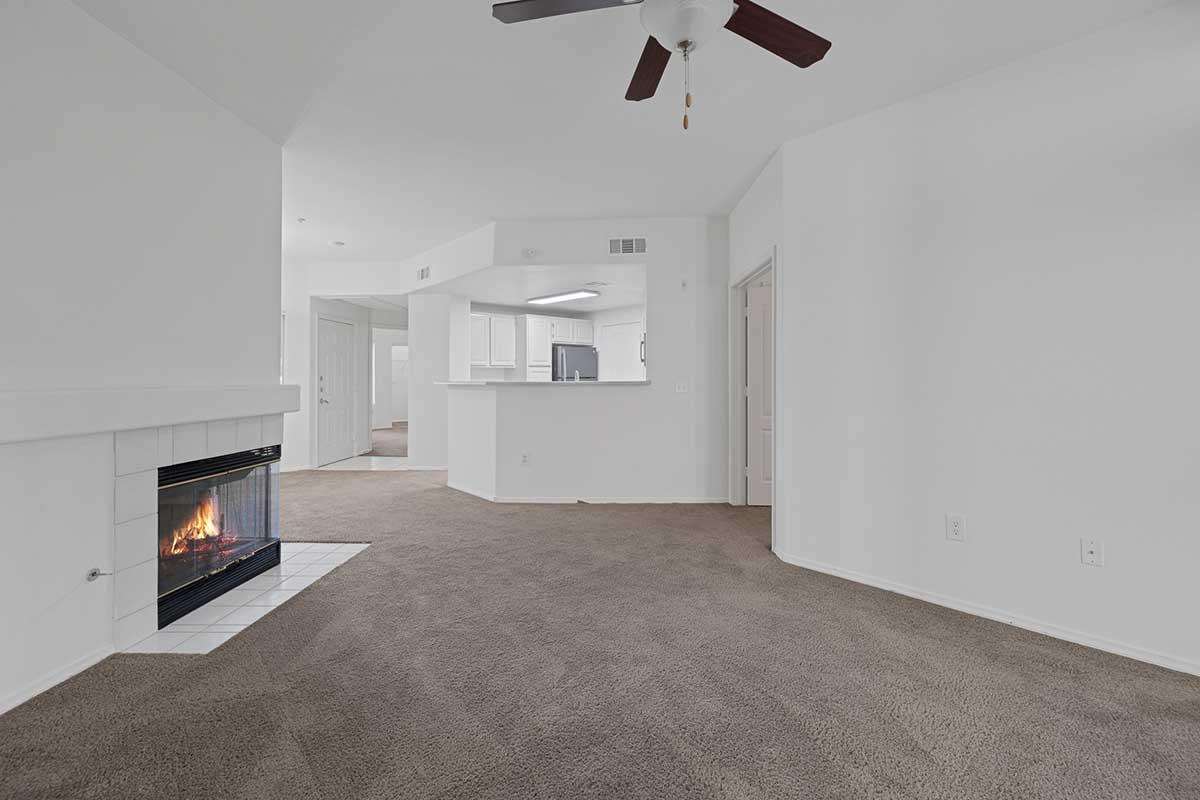 A spacious and well-lit living area featuring a fireplace, brown carpet, and a ceiling fan. The room has white walls and an open layout leading to a kitchen area, with a doorway visible in the background.