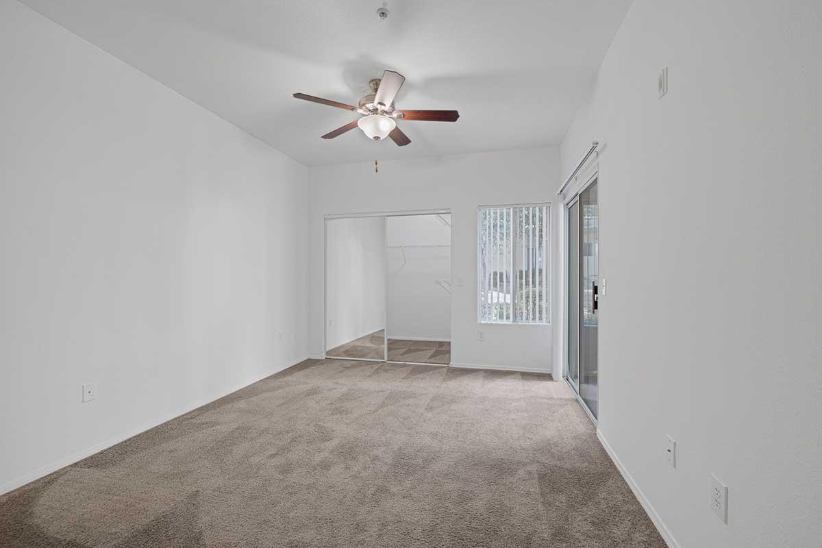 A spacious room with light beige carpet, featuring a ceiling fan with wooden blades. The walls are painted white, and there is a large sliding glass door leading to an outdoor area. Adjacent to the door, there are sliding mirror closet doors. The room has a bright, open feel with natural light coming in.