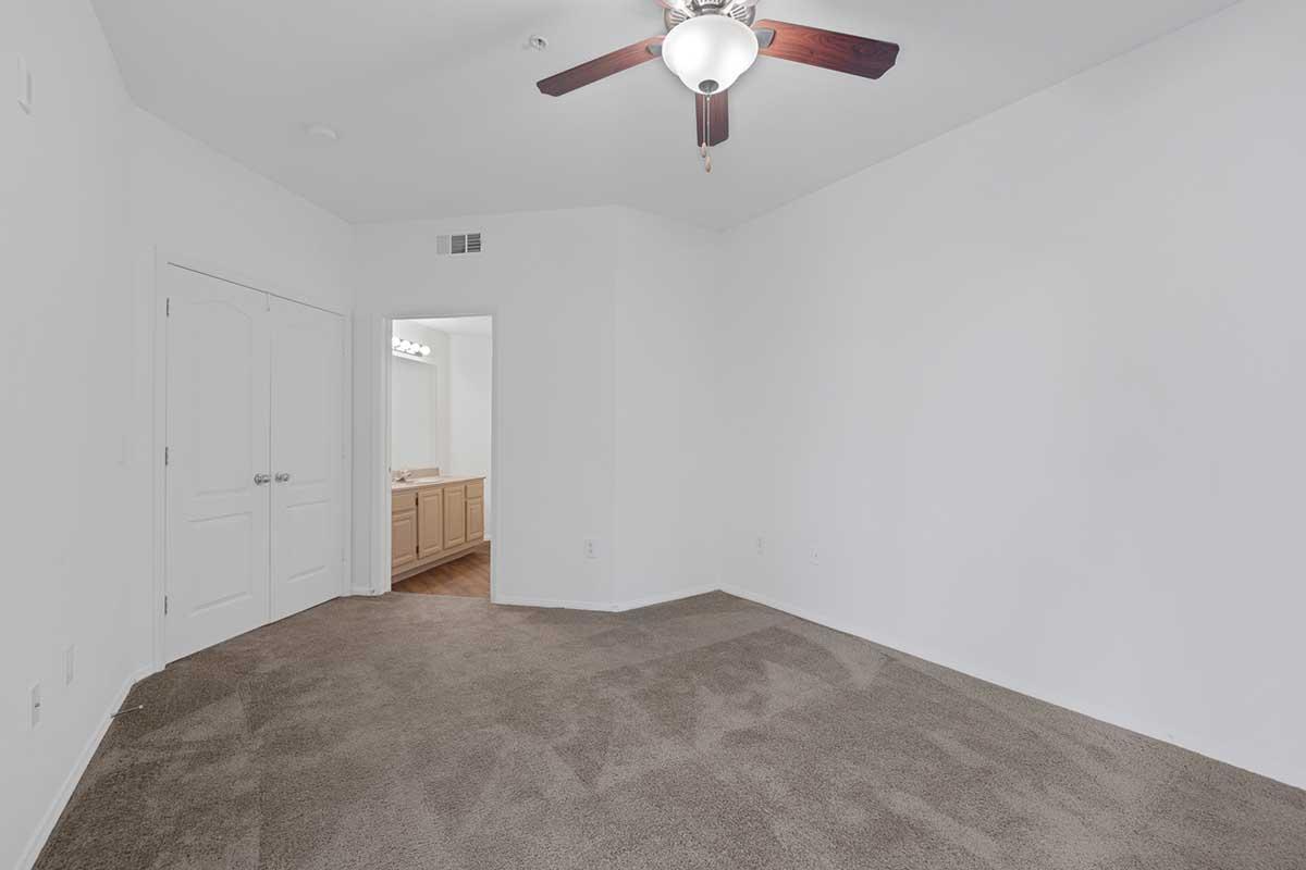 A spacious, empty room with light-colored walls and a ceiling fan. The floor is carpeted in a neutral shade. To the left, there are double doors leading to another area, while a doorway on the right leads to a bathroom with wooden cabinets visible in the background.