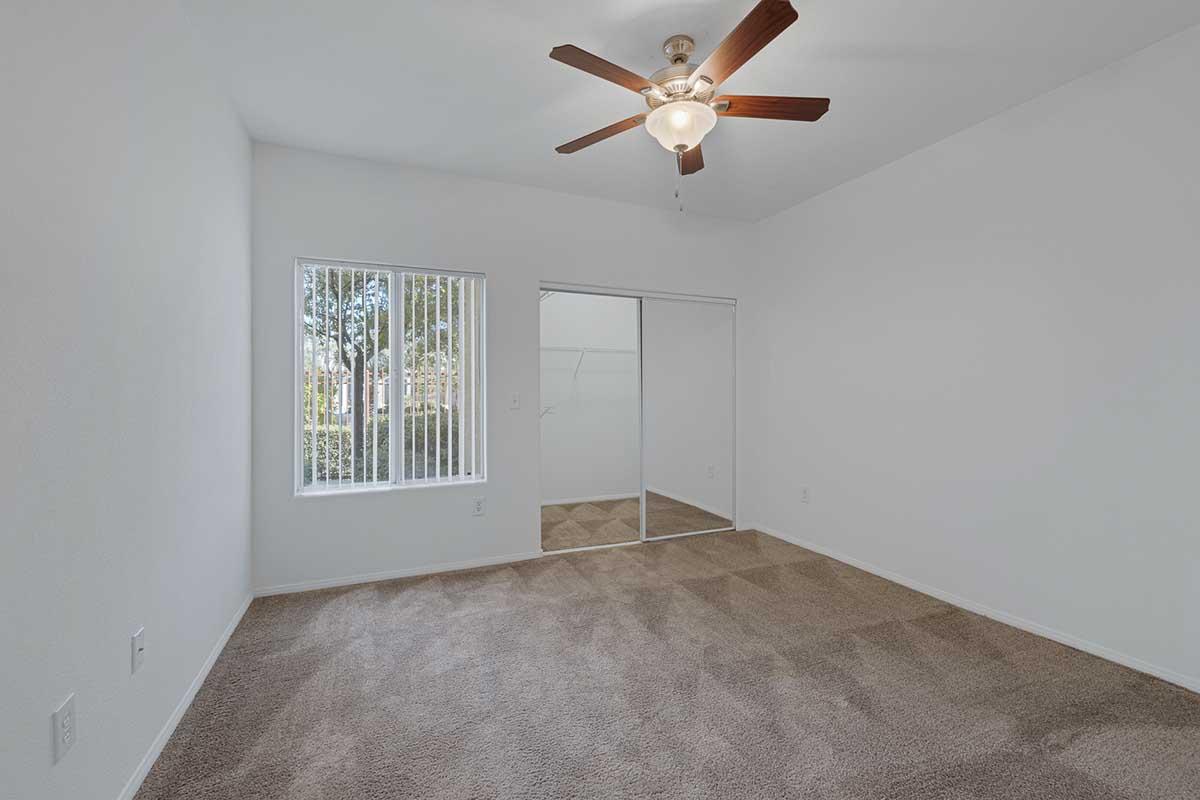 A spacious, empty bedroom with light beige carpet, a ceiling fan, and white walls. There’s a window with vertical blinds allowing natural light, and a closet with mirrored sliding doors. The room is uncluttered, providing a fresh and airy atmosphere.