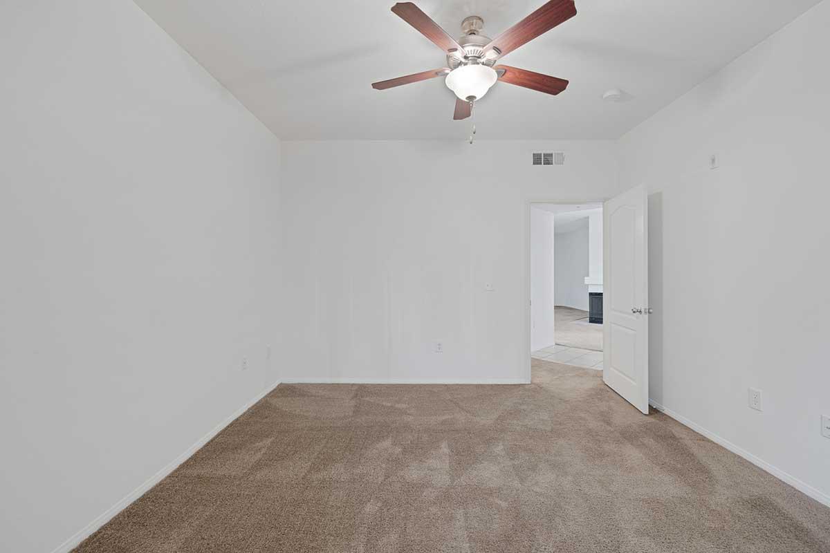 A vacant room featuring light-colored walls and a tan carpet. There is a ceiling fan with a light fixture, and a door is slightly ajar on the right. The room appears spacious and uncluttered, with natural light potentially coming from an adjacent area.