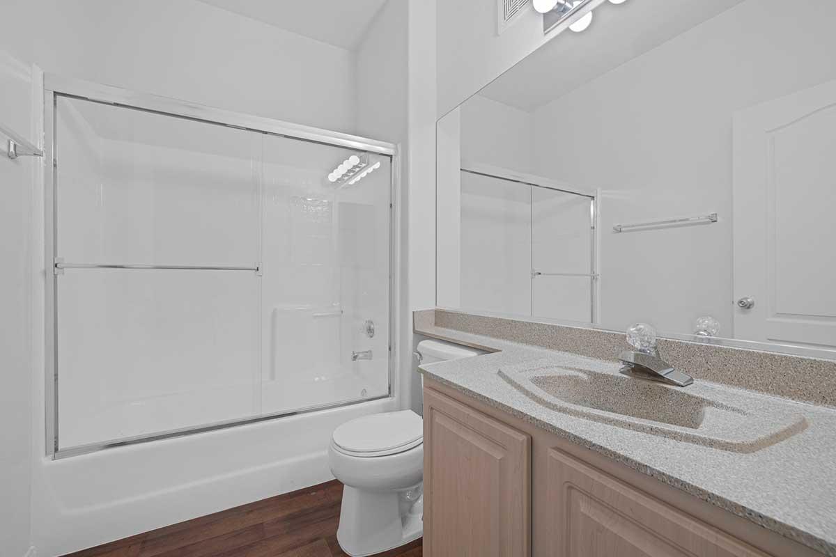 A modern bathroom featuring a clear glass shower enclosure, a large mirror above a countertop with a sink, and a toilet. The walls are painted in a light color, and the floor is made of wooden planks, creating a clean and spacious atmosphere.