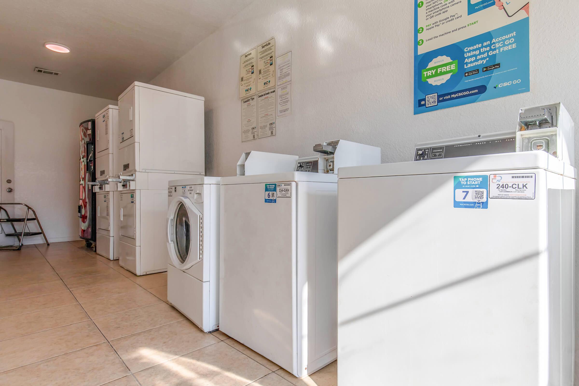 A clean laundry room featuring several white washing machines and dryers lined up against a wall, with informational posters on the nearby wall. The floor is tiled, and there is a small set of stairs in the corner.