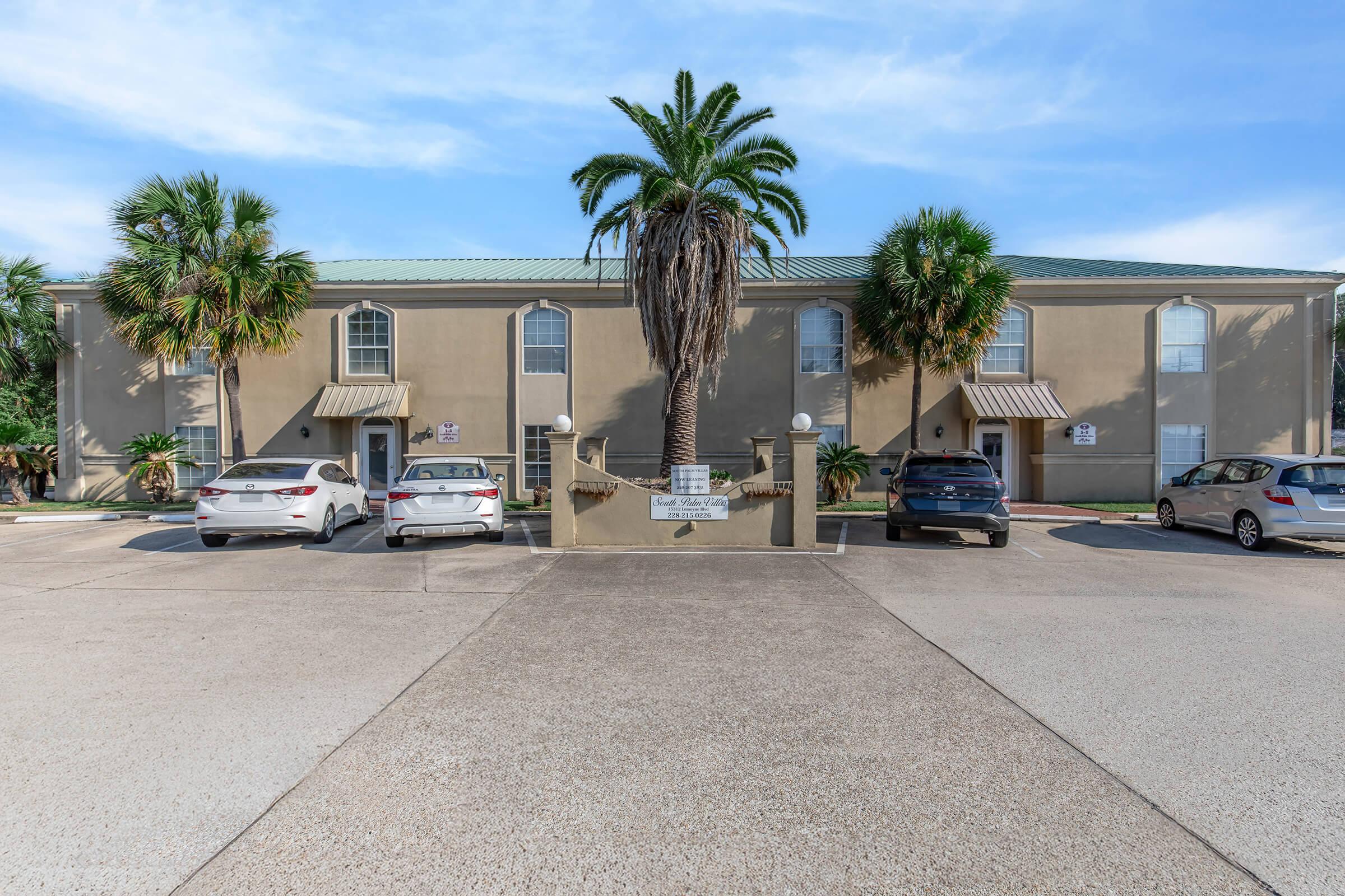 A parking area in front of a two-story residential building with palm trees. Several cars are parked, and there are signs visible near the entrance. The sky is clear with some clouds, creating a bright atmosphere.
