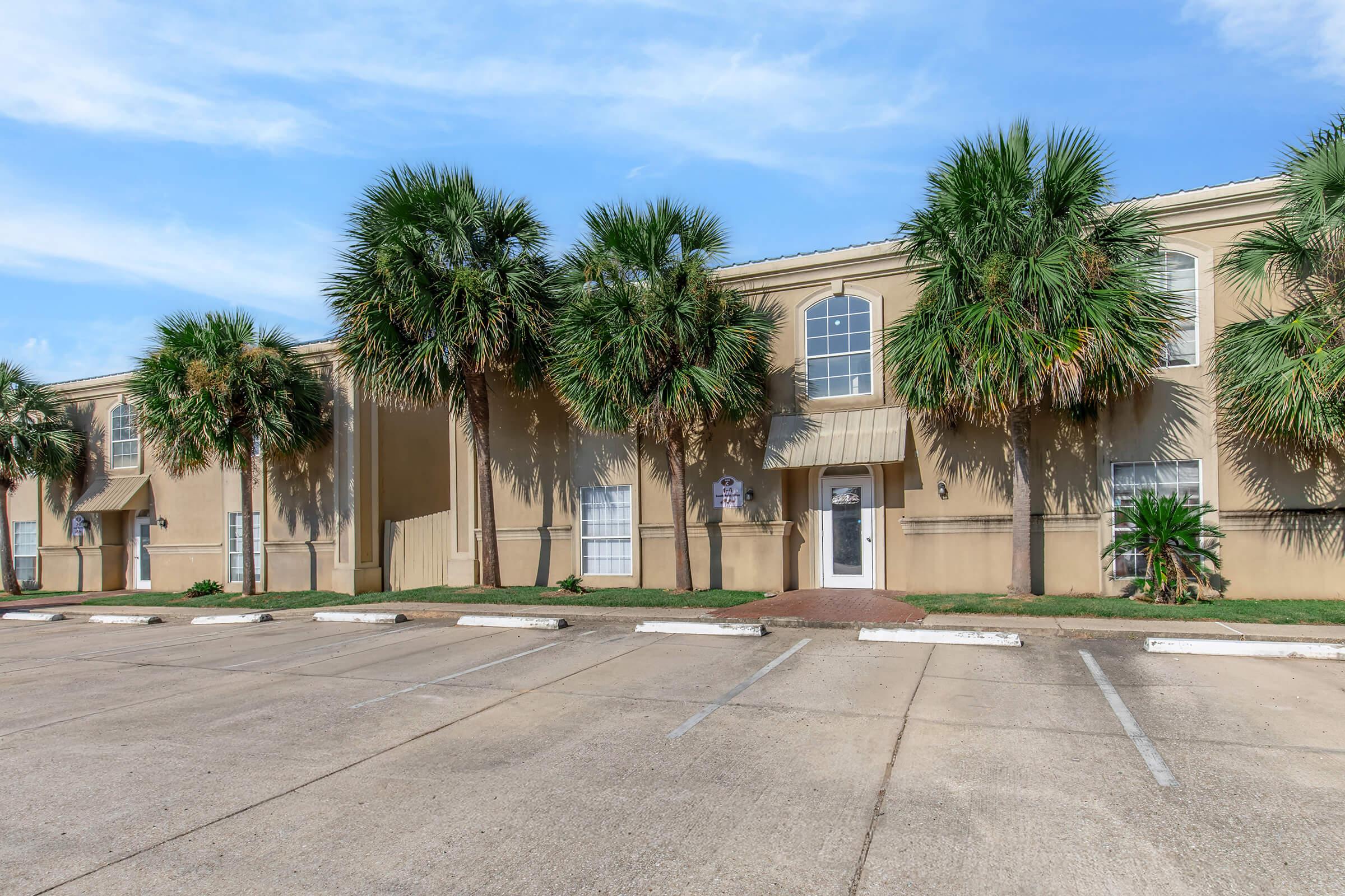 A beige building with multiple windows, bordered by palm trees. The front area features a well-maintained lawn and a paved parking lot with marked spaces. The sky is blue with some clouds, creating a bright, inviting atmosphere.