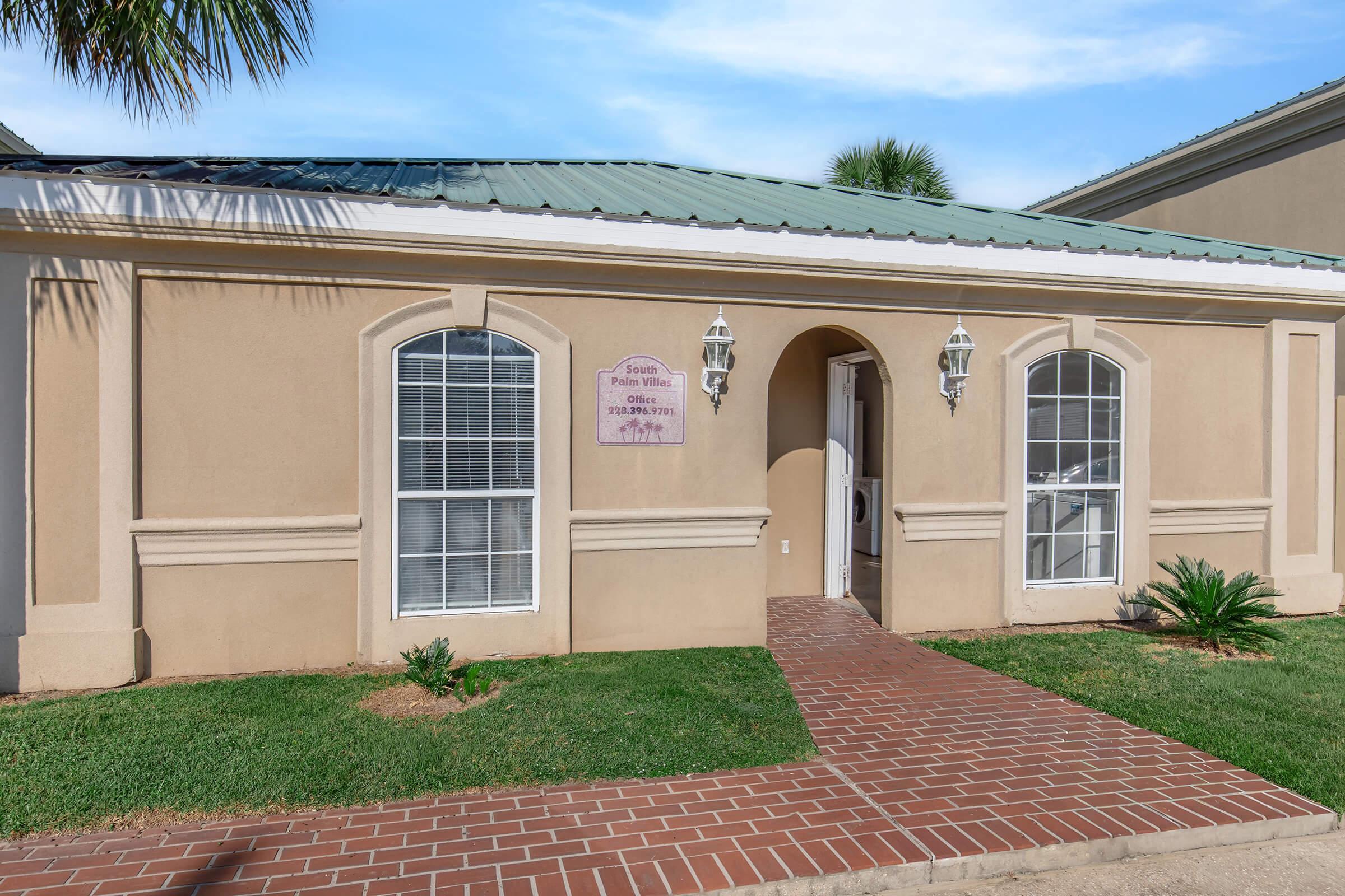 A beige building with a green metal roof, featuring two large arched windows and two lantern-style light fixtures next to the entrance. A brick pathway leads to the door, and there are small shrubs and palm plants in the grass. A sign on the wall reads "Facility Name."