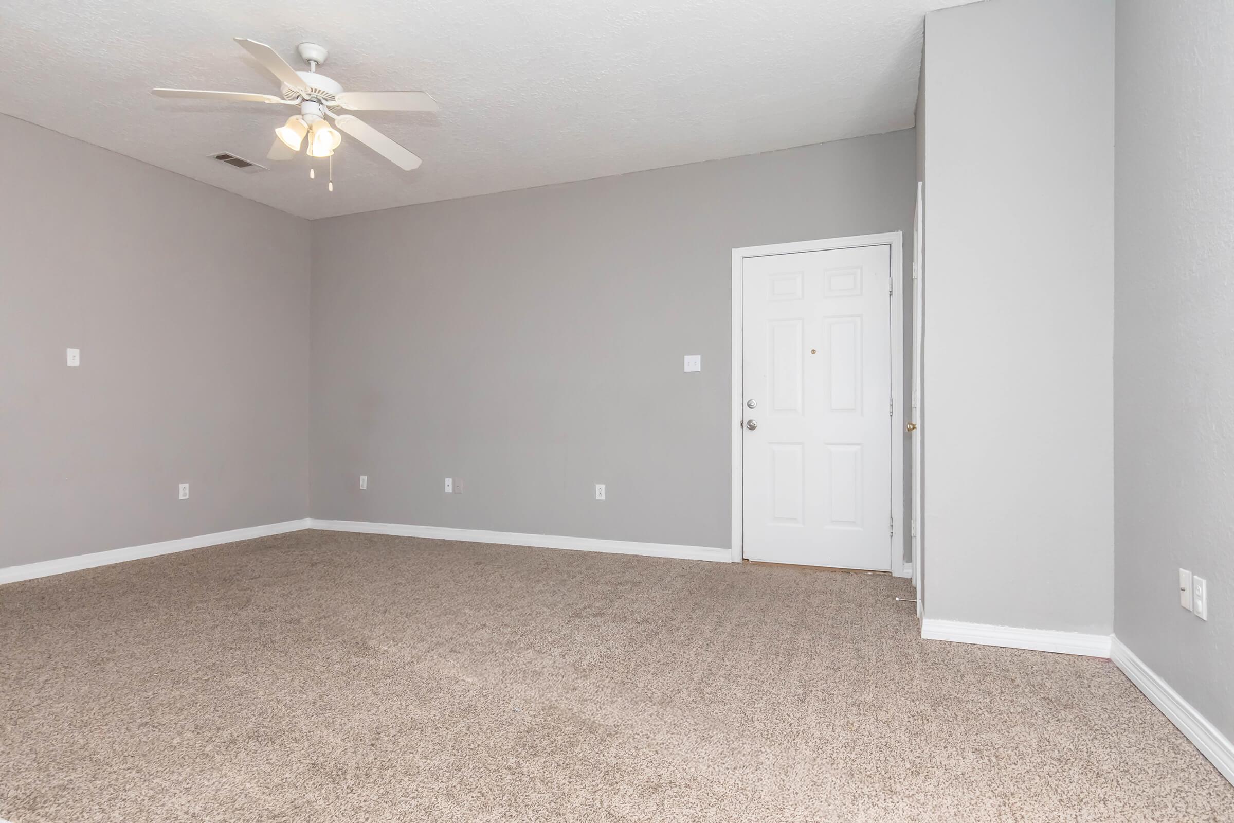 Empty room with light gray walls and beige carpet. A ceiling fan is mounted on the ceiling, and a white door is visible on the right side of the image. There are no furnishings or decorations, giving the space a minimalist appearance.