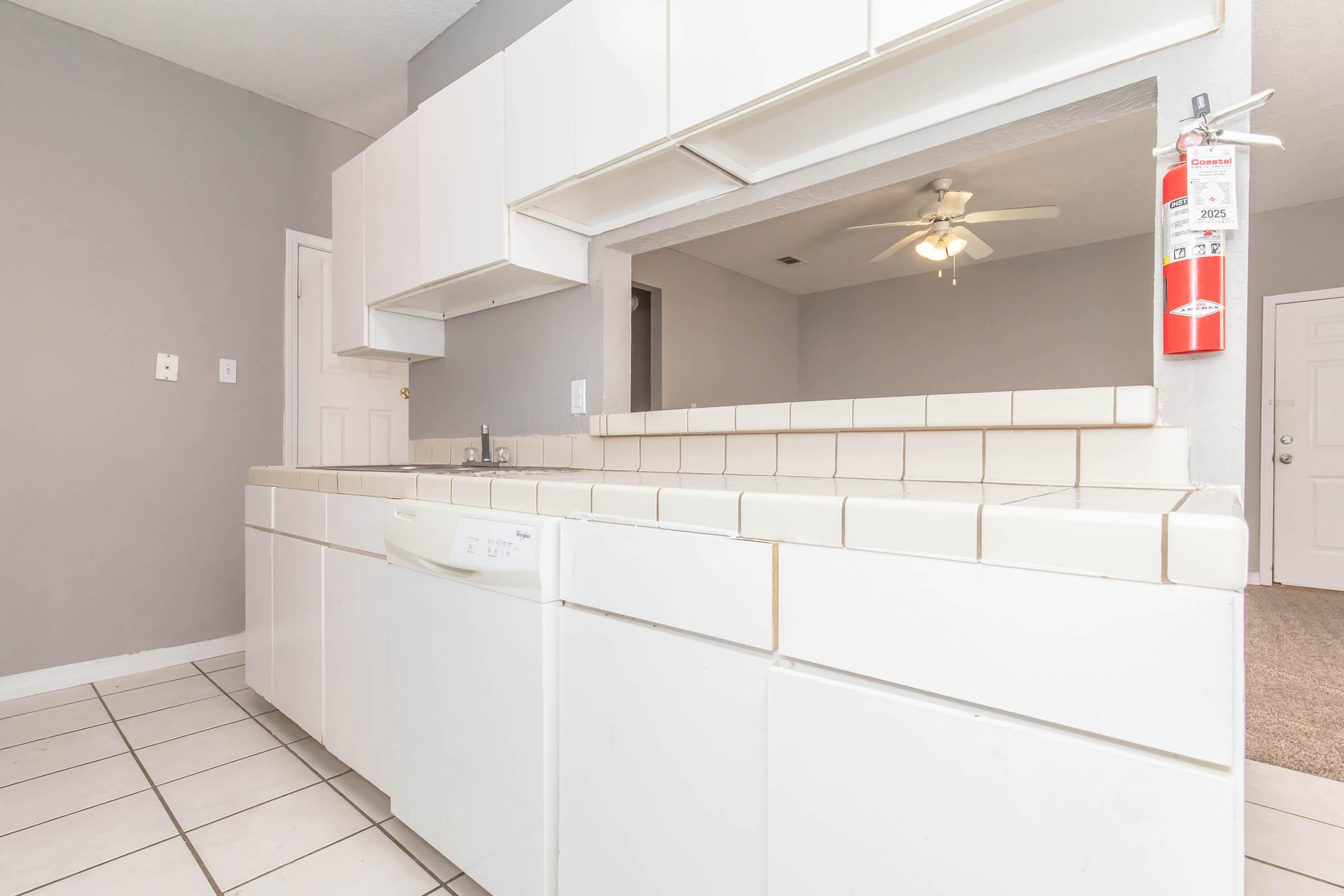 A view of a kitchen featuring white cabinets, a countertop with tiled surface, a dishwasher, and a light ceiling fan. The walls are painted gray, and there's a fire extinguisher mounted on the wall. The space opens into a living area with carpet flooring and a door leading outside.